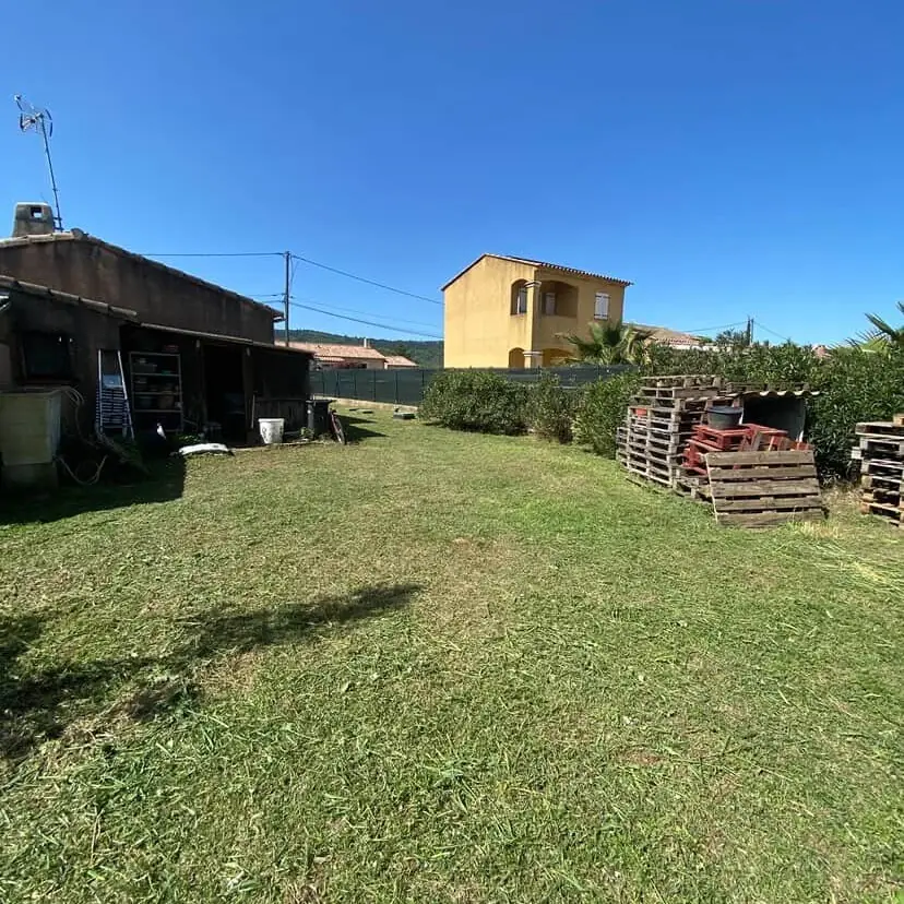 Pelouse tondue devant un cabanon et des palettes, maison ocre et ciel bleu, à Pourrières