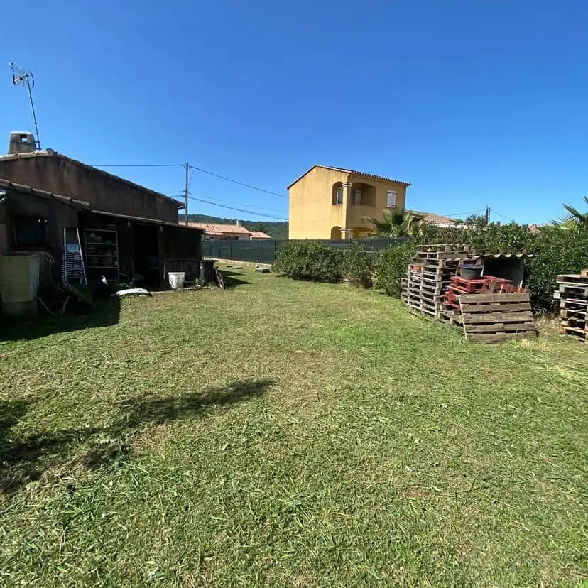 Pelouse tondue devant un cabanon et une maison ocre, ciel dégagé